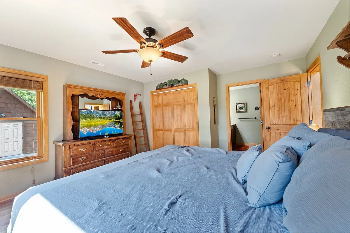 A comfortable bedroom with a bed dressed in blue bedding, a wooden dresser topped with a TV, and a ceiling fan overhead. The atmosphere is serene and cozy, perfect for unwinding with a favorite show or relaxing after a day in the mountains. Sunlight streams softly into the room, enhancing the sense of peace.