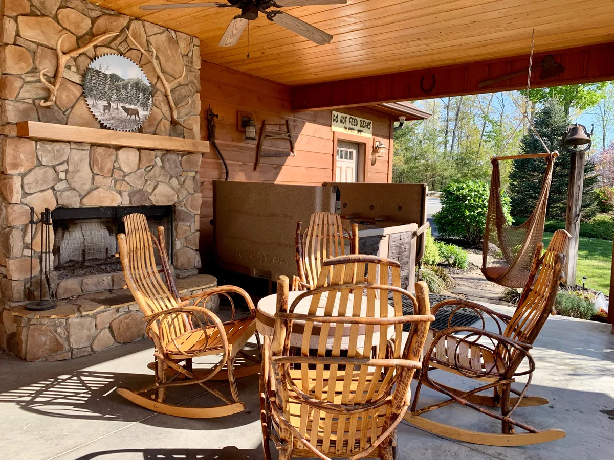 A cozy covered deck with a stone outdoor fireplace, wooden rocking chairs, and a hammock swing, blending rustic comfort with inviting social spaces. The setting is surrounded by nature, perfect for both relaxation and conversation.