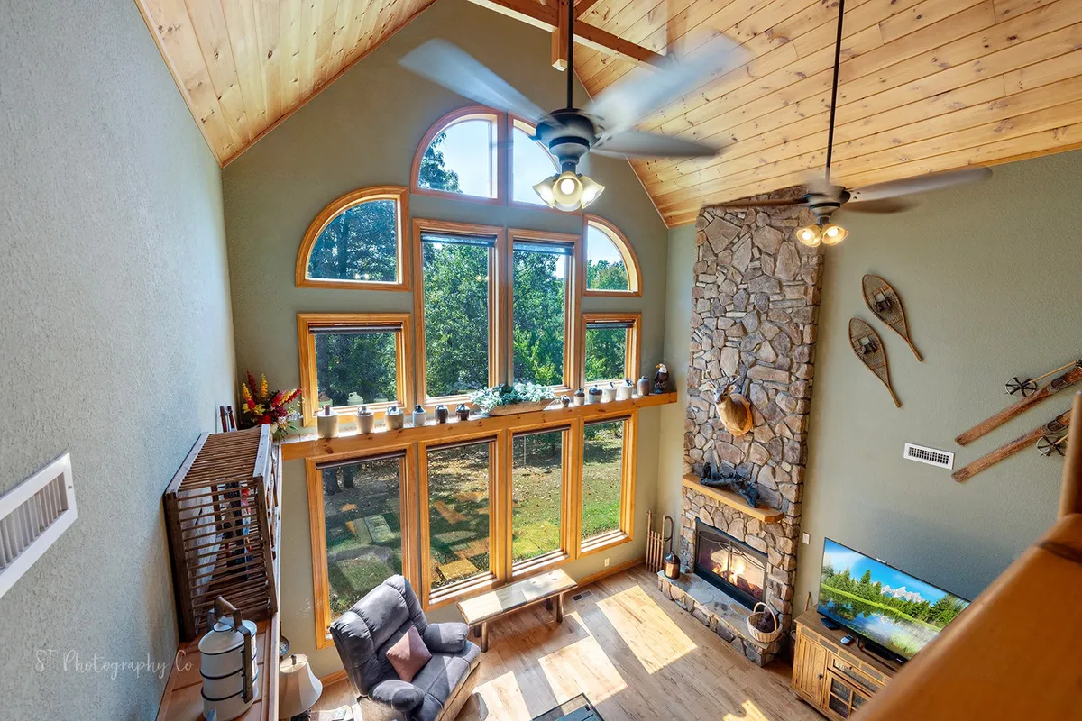 This image depicts a spacious living room featuring a stone fireplace, comfortable seating, large windows, and a wooden ceiling. Natural light pours in, creating a warm, inviting atmosphere ideal for gathering and socializing.