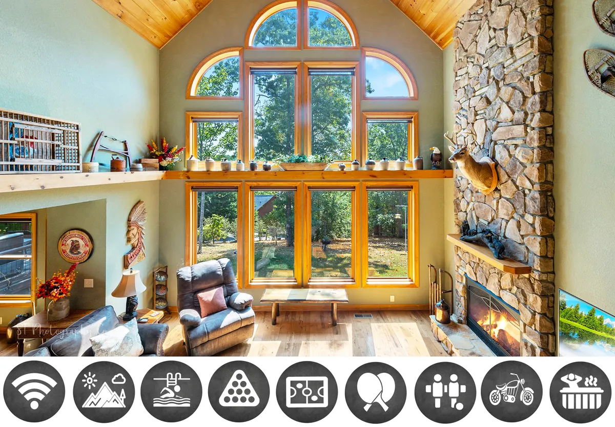 The image showcases a warm and inviting cabin interior with soaring wooden beams, a stunning stone fireplace, oversized windows flooding the space with natural light, and plush seating arranged for relaxation. The sunlight pours in, illuminating the rustic charm, while the view outside hints at the serene beauty of the Smoky Mountains.