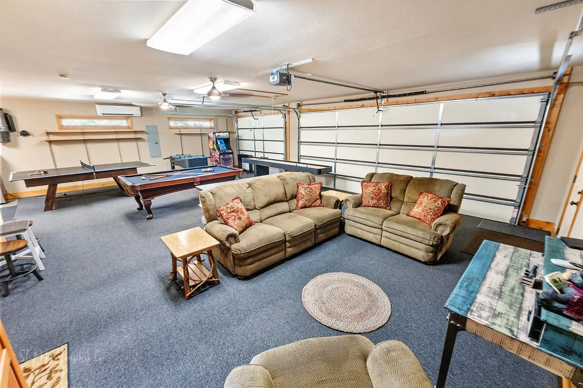 A spacious, multi-purpose game room featuring sofas with decorative pillows, a pool table, a ping pong table, and a garage door. The layout is perfect for group activities, game nights, and relaxed celebrations, offering plenty of room for friends to play and socialize together indoors.