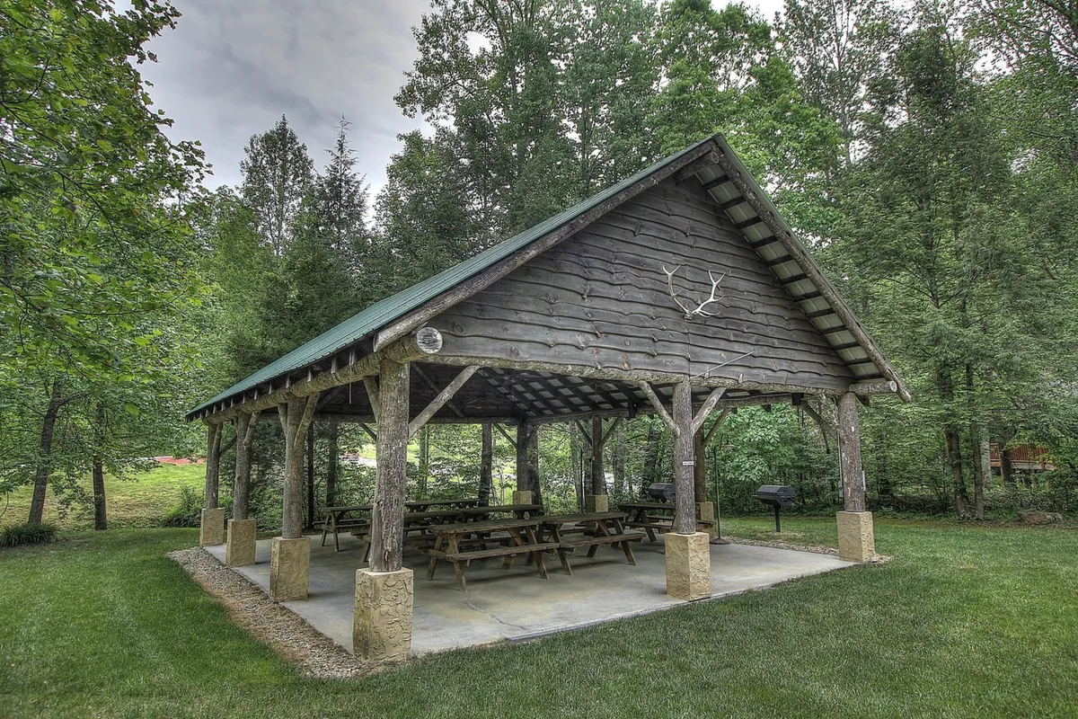 A wooden pavilion with picnic tables and a grill area sits amidst lush greenery, providing an ideal gathering place for outdoor meals, barbecues, and group celebrations. The communal space is designed for family picnics and shared experiences in nature.