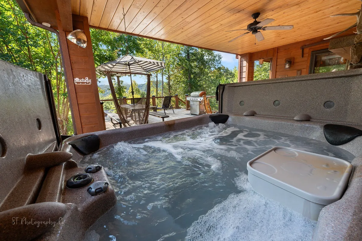 A tranquil outdoor deck featuring a bubbling hot tub, comfy outdoor seating with chairs and an umbrella, and a BBQ grill. Surrounded by greenery, the space is ideal for relaxation and social gatherings, capturing the essence of private mountain living.