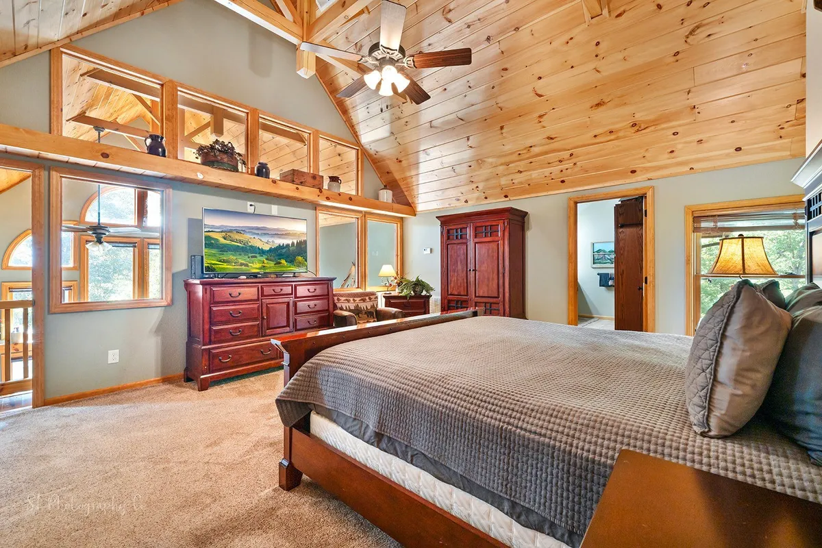 A tranquil bedroom featuring a large, comfortable bed with plush bedding, a wooden ceiling, ample dresser space, and large windows that let in natural light. A landscape painting adds a touch of nature-inspired decor. The room is perfect for relaxing, reading, or watching television, while the windows offer serene nature views.