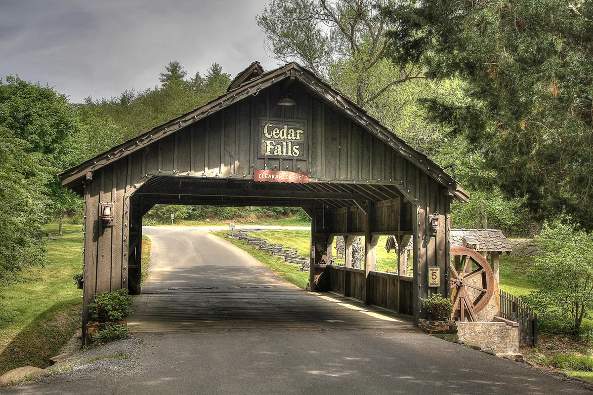 This exterior image features a charming covered bridge, the Cedar Falls sign, a classic water wheel, and lush greenery that welcomes guests into the resort area. The scene encourages outdoor exploration and sets a tranquil mood for nature lovers.