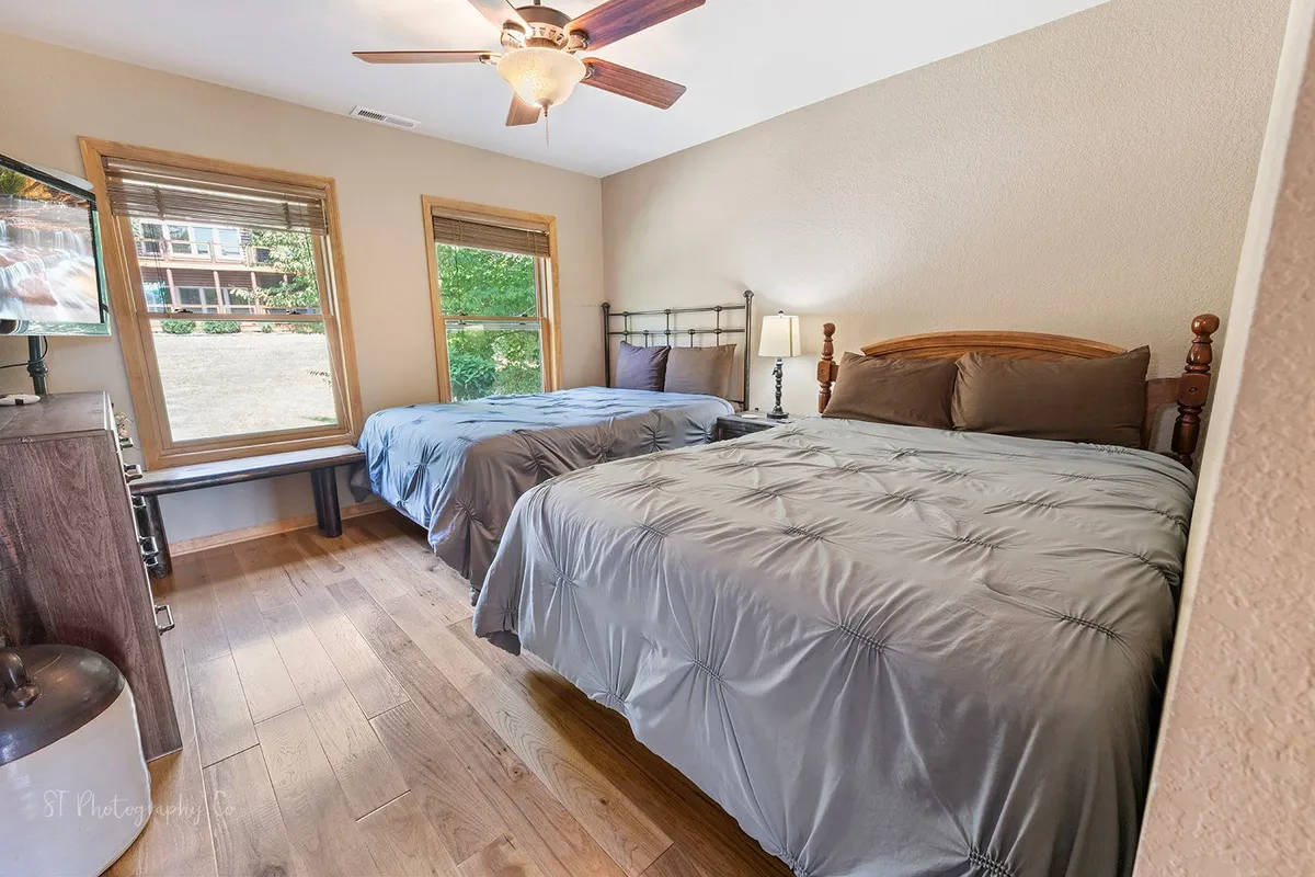 A cozy bedroom with two queen beds dressed in soft gray bedding, a large window offering a scenic view, and a ceiling fan above. The room is inviting and suitable for sharing, and the large window provides a bright, airy atmosphere, ideal for reading or enjoying the landscape.