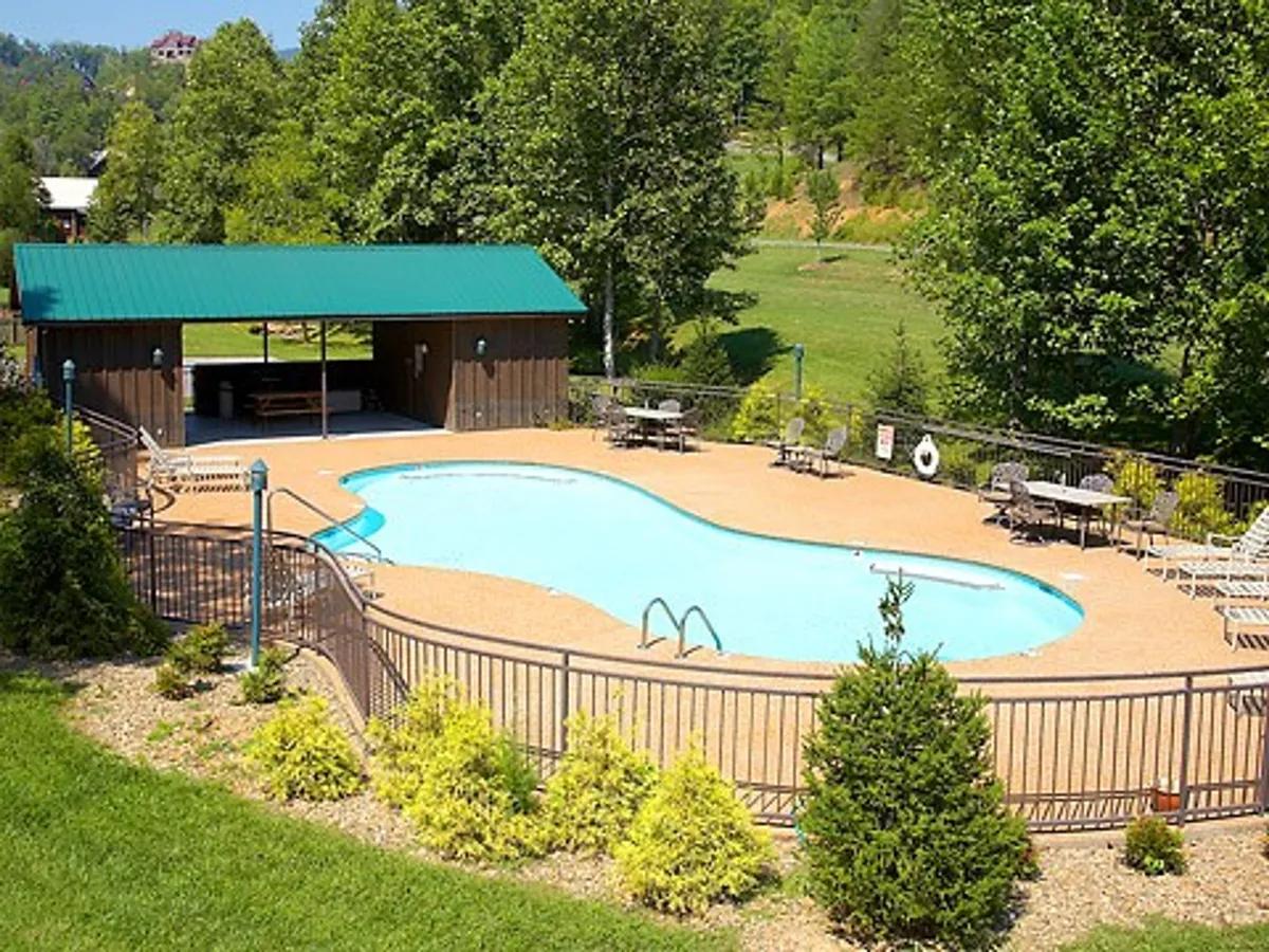 A resort-style swimming pool area with a green-roofed pavilion, ample lounge chairs, and lush trees surrounding the crystal-clear water. The scene captures families enjoying the poolside and the communal resort vibe.