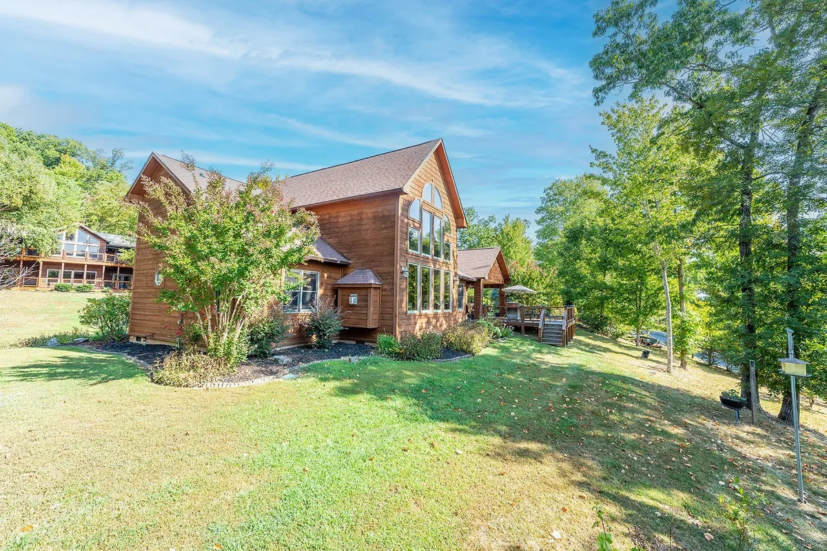 This image shows the exterior of the cabin, featuring large windows, a wooden facade, manicured yard, and surrounding trees. The natural light filters through the trees, highlighting the welcoming and tranquil setting.